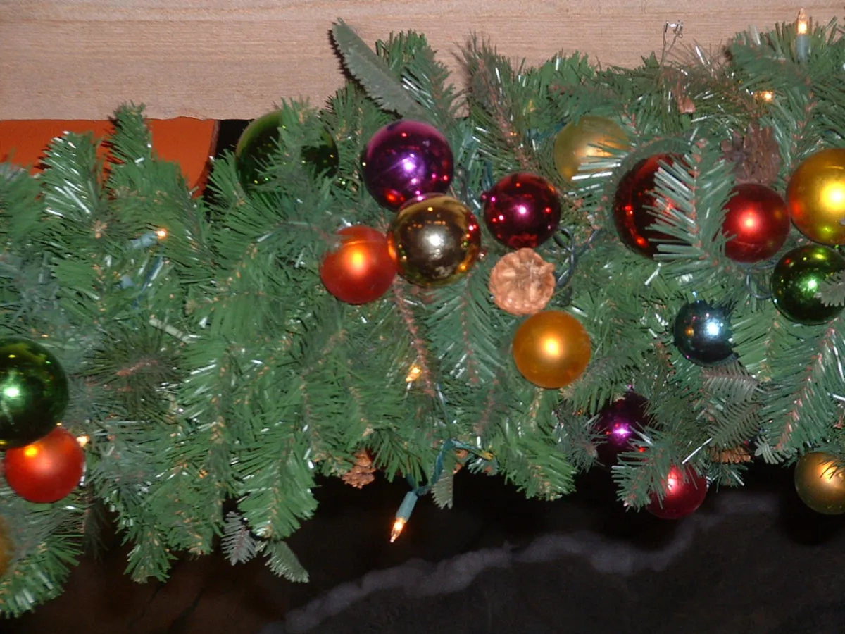 Groene kerstslinger versierd met kleurrijke glazen kerstballen en lichtjes tegen een houten wand.